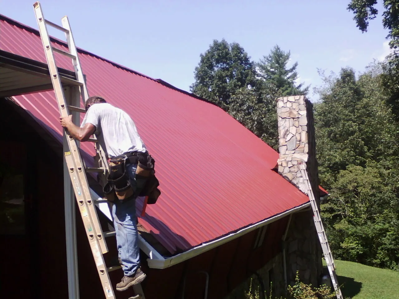 Roofer on a ladder beside a red metal roof and stone chimney — Soffit & Fascia Repair by Roofing Brothers in Hot Springs National Park